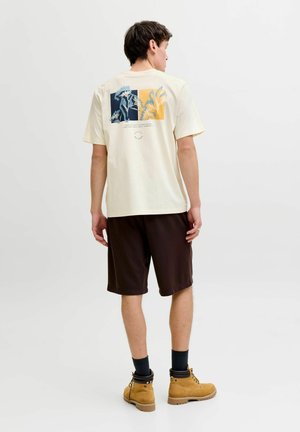 Mann trägt ein beiges T-Shirt mit Grafik, schwarze Shorts, schwarze Socken und beige Stiefel, steht mit dem Rücken zur Kamera vor weißem Hintergrund.