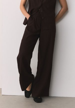 Persona che indossa pantaloni neri larghi e un gilet senza maniche abbinato, in piedi con le mani nelle tasche contro un muro bianco.