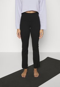 Pantalons de yoga noirs avec une taille ajustée et un bas évasé. Associés à un haut court à manches longues lavande, debout sur un tapis noir.