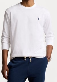 Camicia bianca a maniche lunghe in cotone con collo a girocollo e un piccolo logo blu navy sul petto, abbinata a pantaloni blu navy e un orologio.