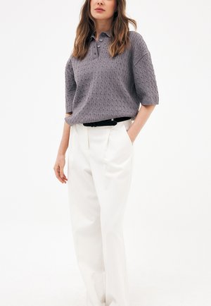 Žena oblečená v fialovom pletenom polokošeli s krátkym rukávom a bielych vysokých nohaviciach s čiernym opaskom, stojaca s jednou rukou v vrecku.