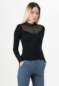 Ung kvinna med blont ombrehår bär en svart långärmad topp med spetsdetaljer och blå byxor, stående med händerna i fickorna.