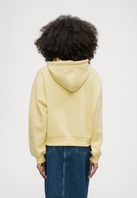 Άτομο με σγουρά μαλλιά, φορώντας ένα ανοιχτό κίτρινο φούτερ και μια μπλε τζιν φούστα, στέκεται με την πλάτη γυρισμένη απέναντι σε έναν απλό φόντο.