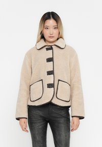 Beige oversized faux shearling jacket with a rounded collar, four black buttons, and contrasting dark trim. Features two front pockets.