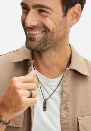 Mannen's zilveren hangerketting met een rechthoekig ontwerp en textuuraccenten. Draagt over een wit T-shirt en een beige jas.