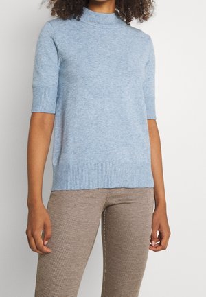 Femme portant un pull à manches courtes bleu clair avec des poignets et un ourlet côtelés, assorti à un pantalon beige à carreaux.
