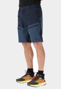 Shorts da esterno blu navy con multiple tasche con zip, vita elastica e pannelli in blu più chiaro a contrasto. Abbinati a sneakers nere e arancioni.