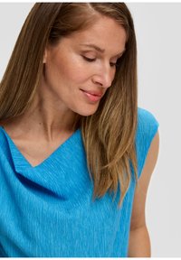 Top blu brillante con scollo drappeggiato e testurizzato. Il tessuto è morbido e leggermente cadente, con maniche corte e una vestibilità comoda.