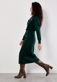 Robe tricotée verte à manches longues, avec deux poches avant, des boutons dorés et une taille cintrée. Portée avec des bottes marron hautes jusqu'aux genoux.
