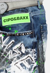 Jean en denim bleu foncé avec des éclaboussures de peinture verte. Doté d'une étiquette vert fluo, de perles noires décoratives et d'un motif graphique stylisé.