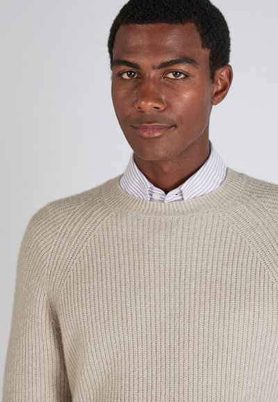 Beige gestrickter Pullover mit Rippstruktur und Rundhalsausschnitt, über einem helllila und weißen gestreiften Hemd getragen.