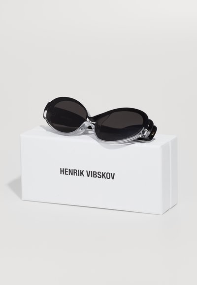 Lielas melnas saulesbrilles ar caurspīdīgām plastmasas detaļām, ar gludu izliekumu, izliktas uz baltas kastes ar uzrakstu "HENRIK VIBSKOV."