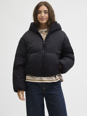 Casaco puffer preto com capuz, apresentando um fecho frontal e um design volumoso. O modelo veste uma camisa às riscas e calças de ganga por baixo.