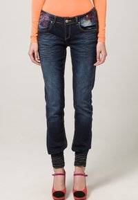 Nainen, jolla on päällään tummansiniset kirjailtujen farkkujen, joissa on rennot hihansuut, oranssi pitkähihainen paita ja punaiset korkokengät mustilla kärjillä.