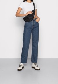 Mujer con una camiseta corta blanca, jeans de tiro alto azul, botas blancas con cordones y un bolso cruzado negro.