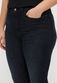Tmavě modré džíny z denimu s středně vysokým pasem, obsahující přední kapsy, zapínání na knoflík a hladkou texturu se jemnými švy.