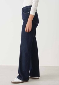 Person wearing dark blue high-waist wide-leg jeans, white long-sleeve top, and white sneakers, standing sideways on a light background.