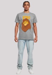 Siva pamučna majica s grafikom lava i natpisom "Kralj" u svijetlo narančastoj boji. Uparena s svijetloplavim trapericama i bijelim tenisicama.