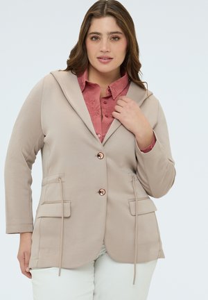 Femme portant un blazer à capuche beige sur un chemisier rose poussiéreux brodé et un pantalon blanc, posant avec la main près du col.
