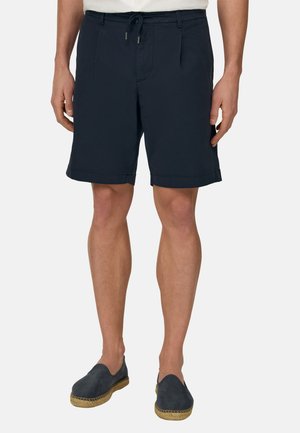 Mann trägt marineblaue Shorts mit Kordelzug und dunkelfarbene Slip-on-Espadrilles, steht vor einem schlichten weißen Hintergrund.