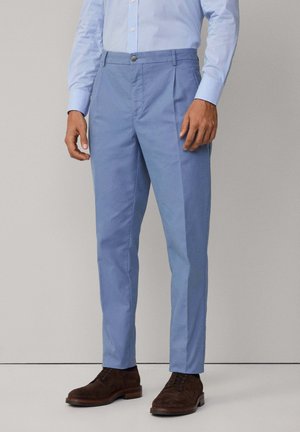 Pantaloni azzurri chiari in morbido cotone, con chiusura a bottoni e pieghe. Scarpe in suede marrone completano l'outfit.