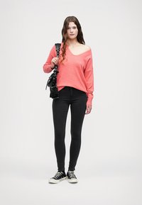 Suéter coral ligero y oversized con escote amplio, combinado con jeans skinny negros y zapatos negros. Modelo sosteniendo un bolso negro pequeño.