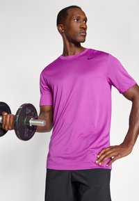 Lila Sport-T-Shirt mit kurzen Ärmeln und rundem Halsausschnitt, mit einem schwarzen Nike-Logo. Das Modell hält in einer Hand ein Gewicht.
