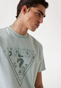Tricou verde menta cu un design grafic triunghiular care include textul "GUESS", accente distresate și mâneci scurte. Material din bumbac.