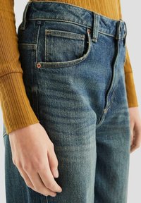 Jeans dżinsowe w odcieniu wyblakłego niebieskiego z wysokim stanem, pięcioma kieszeniami i kontrastowymi przeszyciami; w połączeniu z żółtym swetrem prążkowanym.