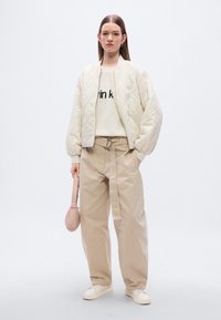 Μπουφάν bomber από κρεμ καπιτονέ πάνω από μπεζ πουλόβερ με μαύρο κείμενο, συνδυασμένο με μπεζ παντελόνι ψηλόμεσο και λευκά αθλητικά παπούτσια, κρατώντας μια ροζ τσάντα.