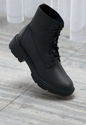 Black leather ankle boot with laces, thick sole, embossed logo on side, displayed on light marble floor against sheer white curtains.