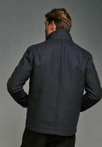 Marineblaues Herringbone-Wollmantel mit hohem Kragen, geradem Schnitt und taillierter Passform. Glatte Textur und dezentes Muster auf dem Stoff.