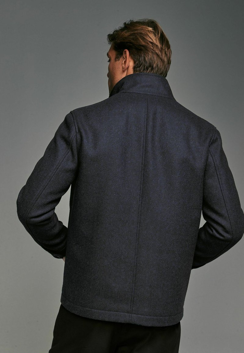 Marineblaues Herringbone-Wollmantel mit hohem Kragen, geradem Schnitt und taillierter Passform. Glatte Textur und dezentes Muster auf dem Stoff.