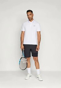 Camisa polo branca com um pequeno logótipo, combinada com calções pretos. O modelo segura uma raquete de ténis, vestindo meias brancas e sapatos de atletismo.