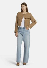 Bruine wol cropped jacket met twee voorzakken, knoopsluiting, gedragen over een witte top en lichtblauwe high-waisted jeans. Zwarte hakken.