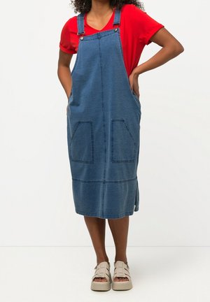 Person iført en rød t-shirt med korte ærmer under en blå denim forklædekjole med forlommer og beige sandaler, stående med hænderne i lommerne.