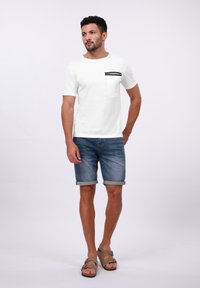 Hvid kortærmet t-shirt med en sort lomme og kant, parret med denimshorts og brune sandaler. Modellen står med den ene hånd på hoften.