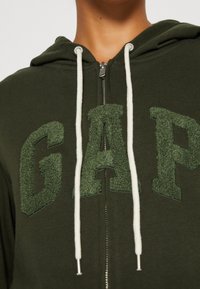 Sudadera con cremallera de color verde oscuro que presenta letras en relieve y textura de "GAP" en un tono a juego, con cordones blancos y un acabado de tela suave.