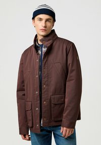 Chaqueta aislante marrón con un cuello alto, botones a presión, dos bolsillos frontales y un acabado texturizado, llevada sobre una camisa de cuadros. Gorro azul marino con rayas.