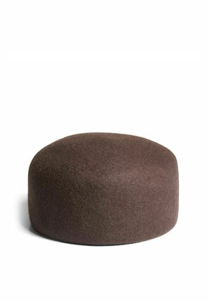 Femme aux cheveux bouclés portant un chapeau marron et un manteau en fausse fourrure gris, les bras croisés, regardant la caméra avec confiance sur un fond uni.