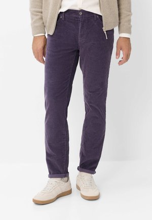 Mand iført lilla kordfløjlsbukser, beige lynlås-sweater, hvid skjorte og hvide sneakers, der står mod en hvid baggrund.