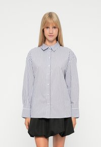 Jonge vrouw met lang blond haar, gekleed in een witte blouse met verticale zwarte strepen en een zwarte rok, staande tegen een witte achtergrond.