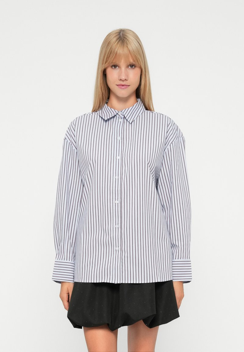 Jonge vrouw met lang blond haar, gekleed in een witte blouse met verticale zwarte strepen en een zwarte rok, staande tegen een witte achtergrond.