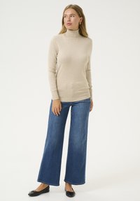 Maglione a collo alto beige in morbido tessuto a maglia, abbinato a jeans in denim blu a gamba larga. Presenta una silhouette aderente e polsini a coste.