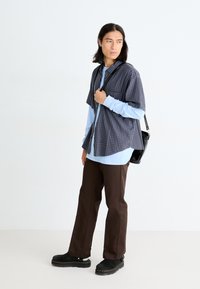 Camisa de cuadros de manga corta en azul marino y rojo sobre una camiseta de manga larga azul claro, combinada con pantalones marrones y zapatos de plataforma negros.