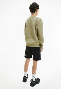 Oliwkowy sweter z długimi rękawami, połączony z czarnymi szortami z dużym białym logo. Widoczne czarne buty sportowe i białe skarpetki.