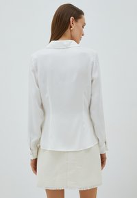 Blusa bianca a maniche lunghe in tessuto lucido e liscio con colletto classico, abbinata a una mini gonna bianca testurizzata con rifiniture in frange.