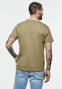 Camiseta de algodón verde oliva con mangas cortas, cuello redondo y un corte relajado, combinada con unos vaqueros de denim azul claro. Tatuajes notables en el brazo.