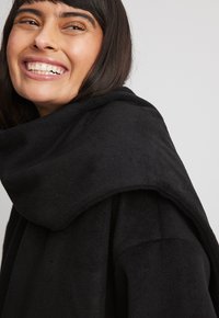 Czarny polarowy strój z szeroką, drapowaną kołnierz. Materiał wydaje się miękki i teksturowany, charakteryzuje się luźnym krojem i minimalnym wykończeniem szwów.