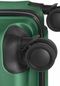 Gros plan sur le coin d'une valise rigide verte avec des roues doubles noires pivotantes et un détail de fermeture éclair.